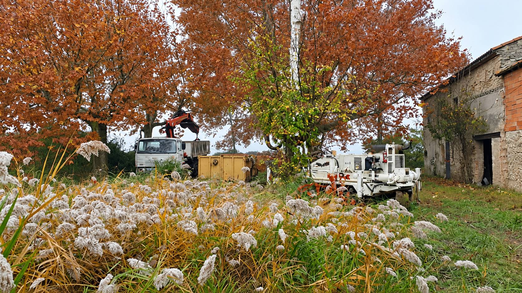 Un camion équipé d’une grue et une machine de forage blanche sont installés dans un jardin brumeux, à côté d’une ancienne maison en pierre et d’arbres aux feuilles d’automne.